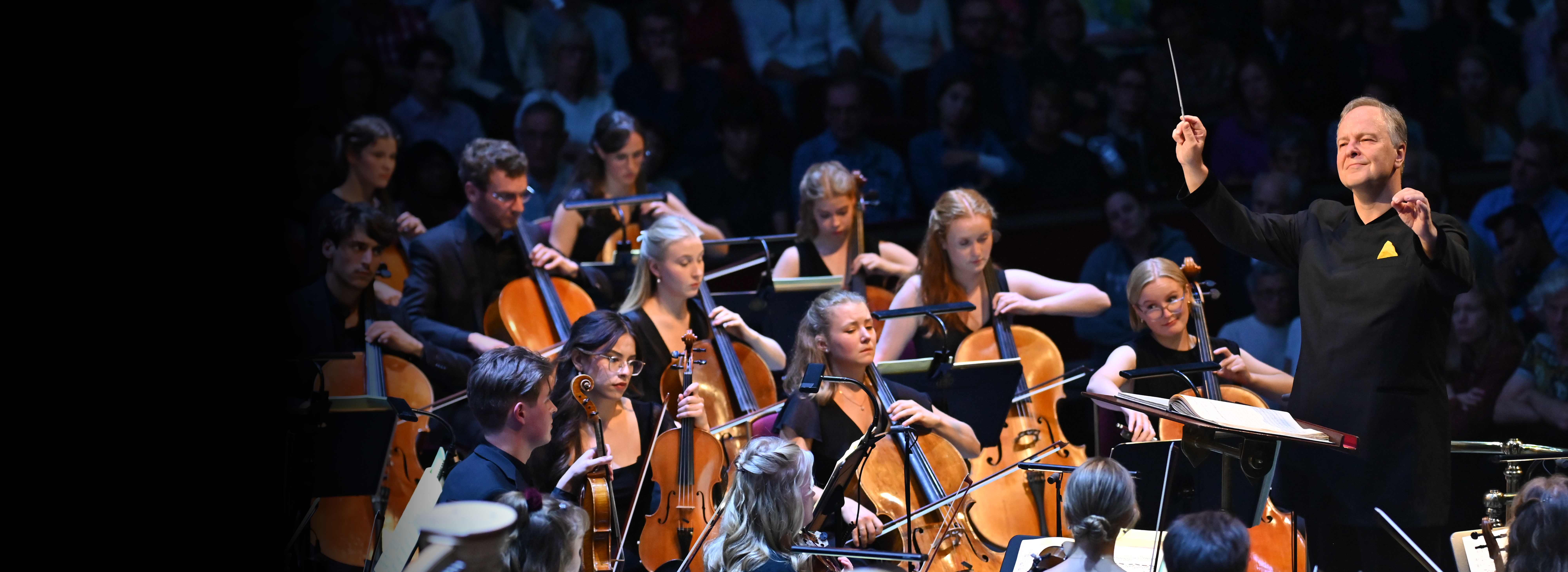 A man conducting a group of musicians, wearing formal black concert attire, playing on string instruments, with an audience in the background.